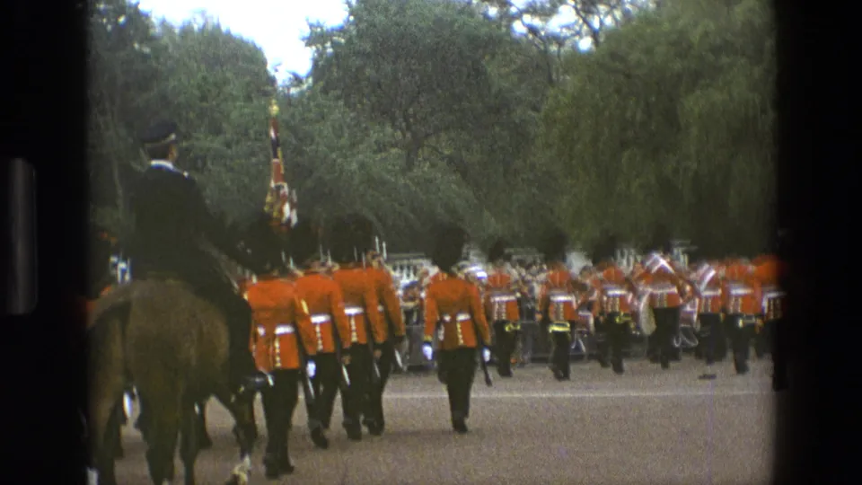 1969 England home-movie frame showing a mounted officer beside a marching group of British guards in red tunics and black hats.