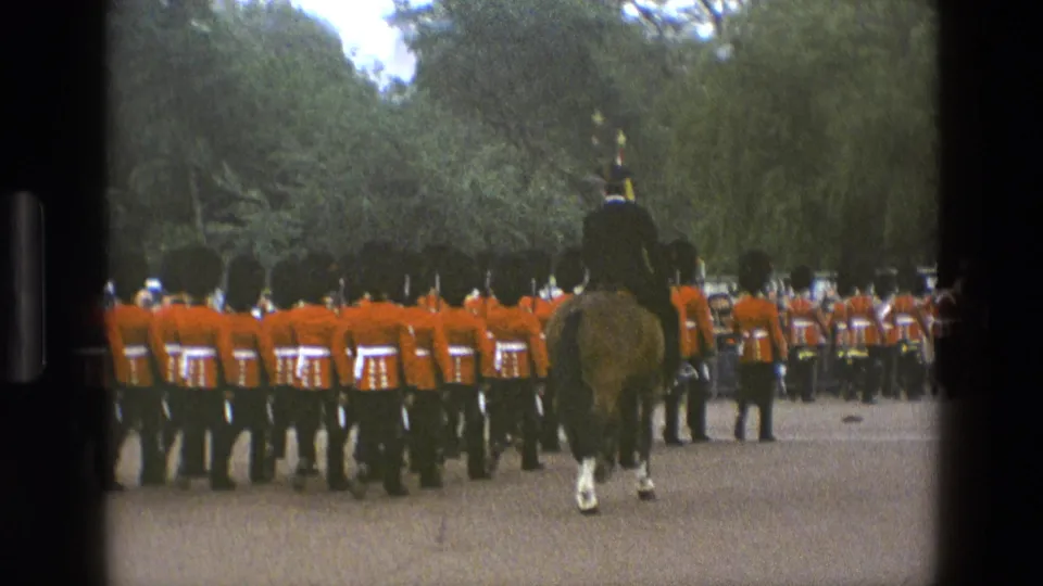 Rear-facing 1969 England home-movie view of ceremonial guards marching in red tunics beneath trees, with a mounted officer ahead.