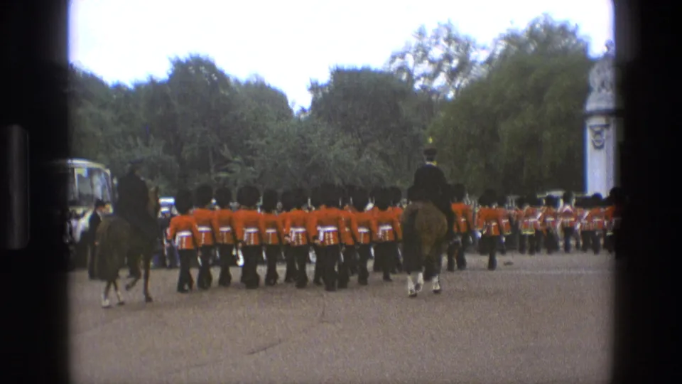 Wide 1969 England parade frame showing guards in red uniforms marching away, mounted riders nearby, trees, road, and a stone structure at right.