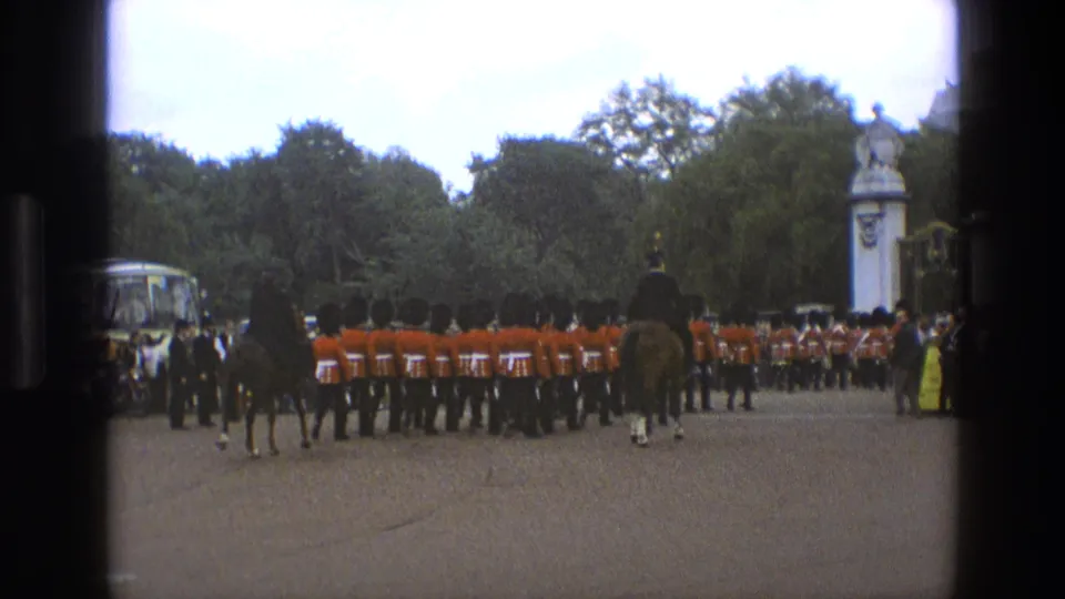 1969 England 8mm frame showing red-coated guards in formation, mounted riders, spectators, trees, and formal gate pillars in a ceremonial setting.