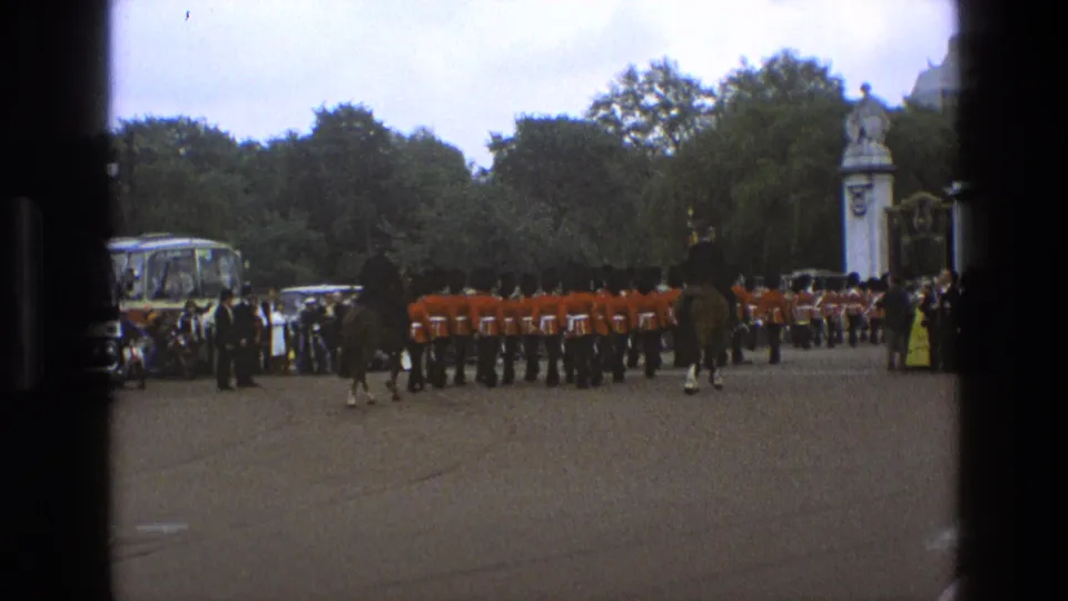 1969 England 8mm frame with guards in red coats, mounted riders, a spectator crowd, trees, formal gates, and a coach in the background.