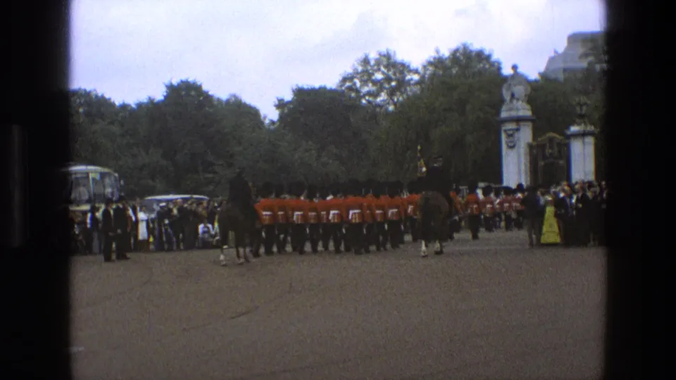 1969 England home-movie frame showing a ceremonial guard line, horseback riders, spectators, trees, formal gate pillars, and a coach nearby.