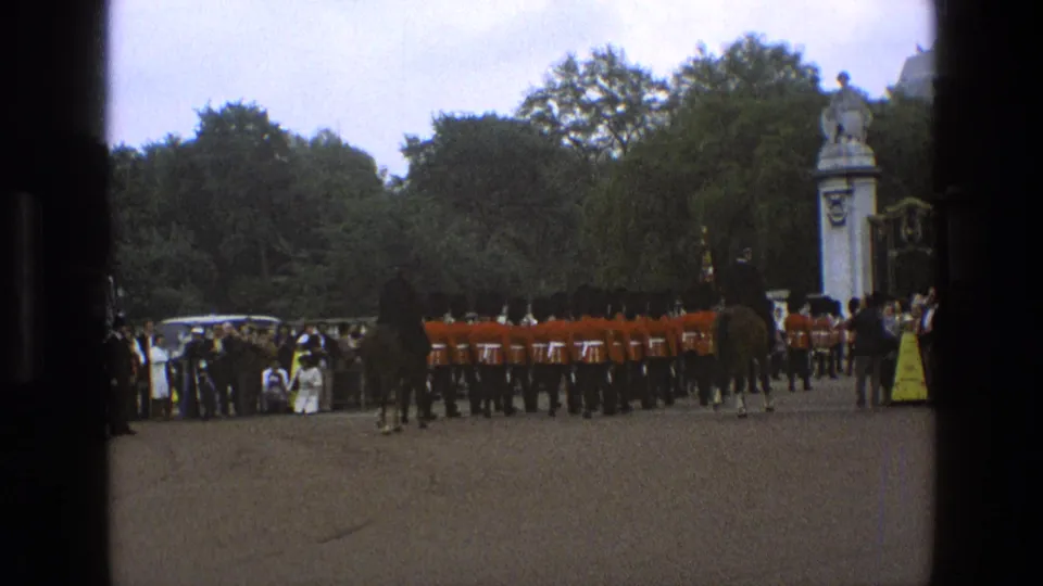 1969 England 8mm frame with mounted guards in the foreground, red-coated soldiers in formation, spectators, trees, and a formal gate backdrop.