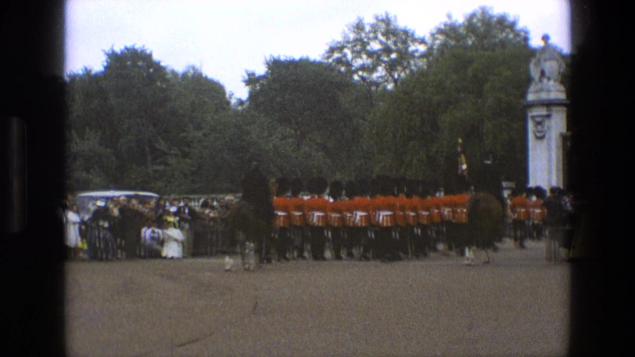 Guards in red tunics march past spectators near stone gate pillars and trees.