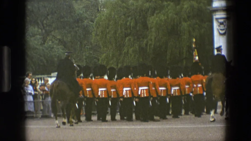 1969 England home-movie frame showing a mounted guard beside foot guards in red tunics moving away across an open ceremonial ground.