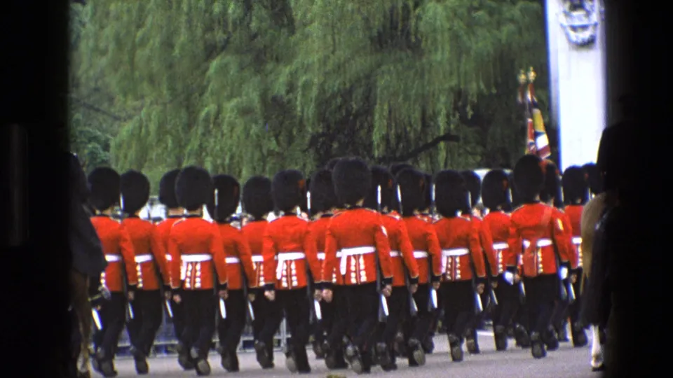 Close 1969 England frame of guards in red tunics and bearskin hats marching away near a ceremonial flag, trees, and a stone gate pillar.