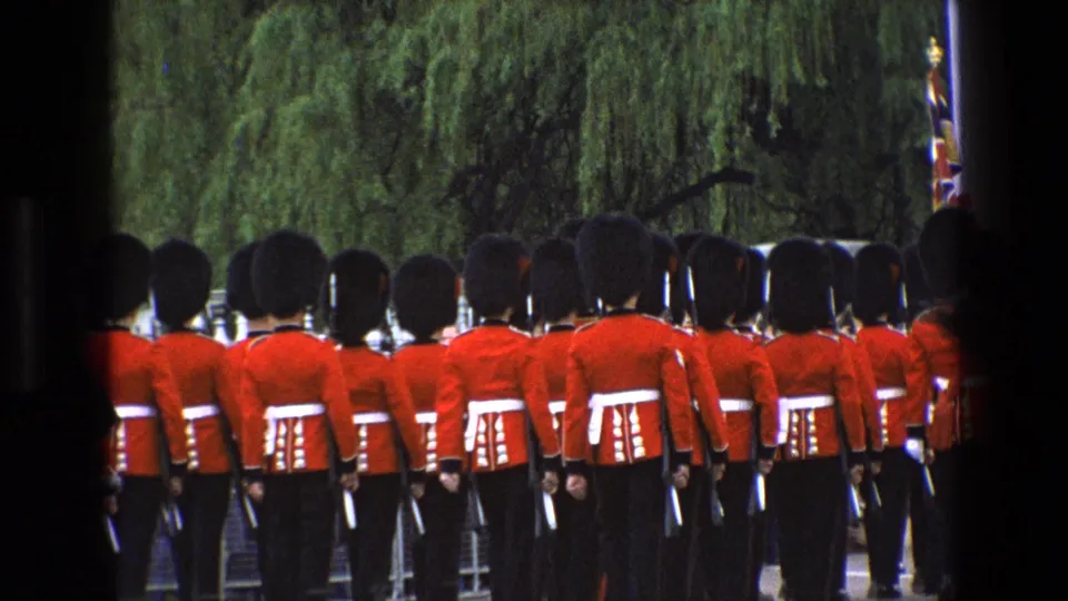 1969 England 8mm frame of ceremonial guards in red tunics and bearskin hats marching past trees behind dark gate bars.