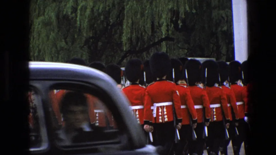 1969 England home-movie frame with red-uniformed guards marching as a black taxi passes across the foreground.