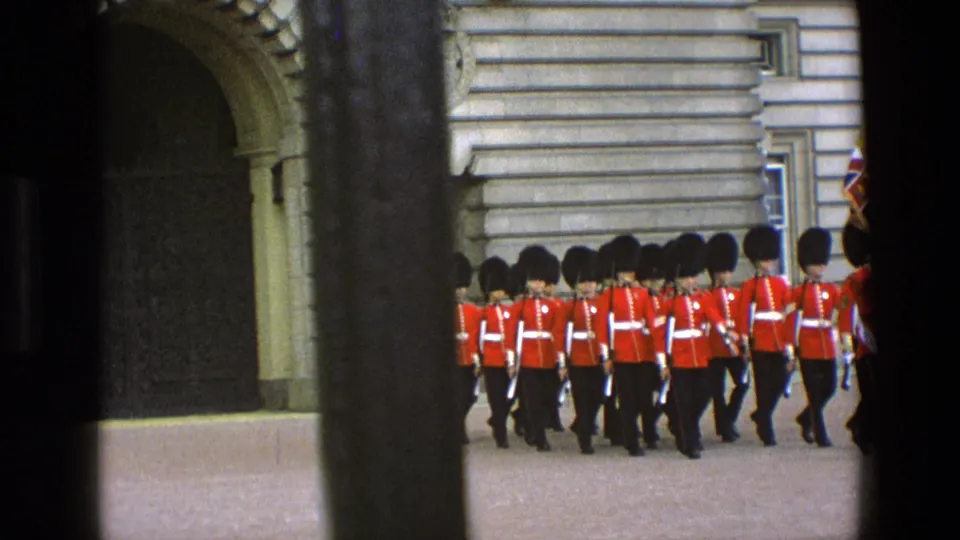 1969 England 8mm frame of ceremonial guards in red tunics and bearskin hats marching beside a stone facade and trees.