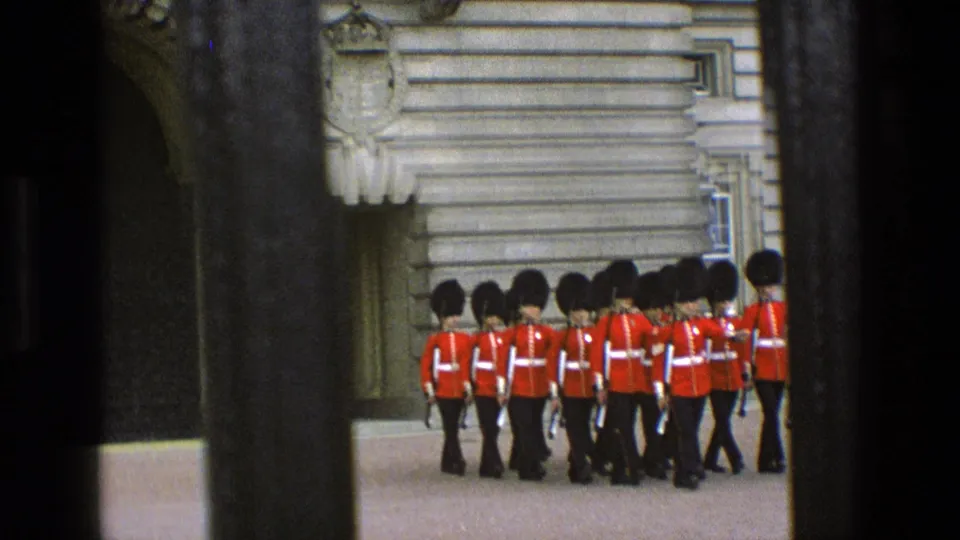 1969 England home-movie frame showing palace guards in red tunics and bearskin hats marching across a courtyard behind railings.
