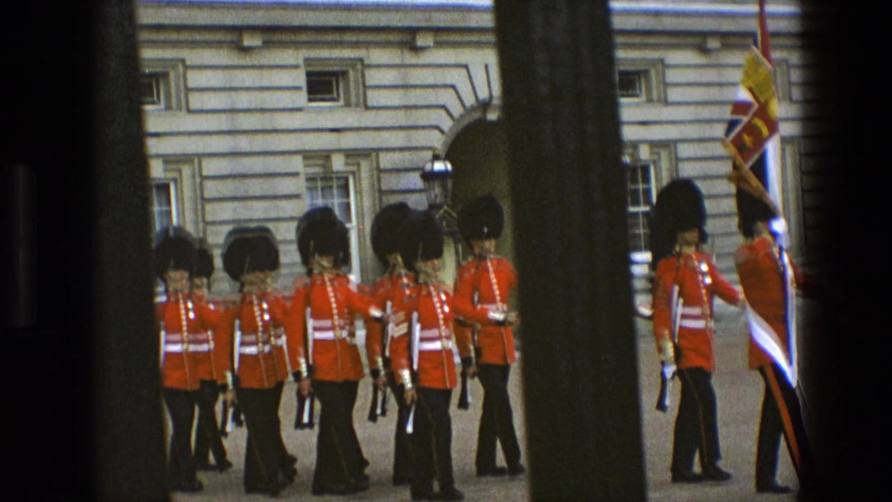 1969 England frame of Buckingham Palace guards marching in red uniforms, seen through vertical gate bars with palace stonework behind them.