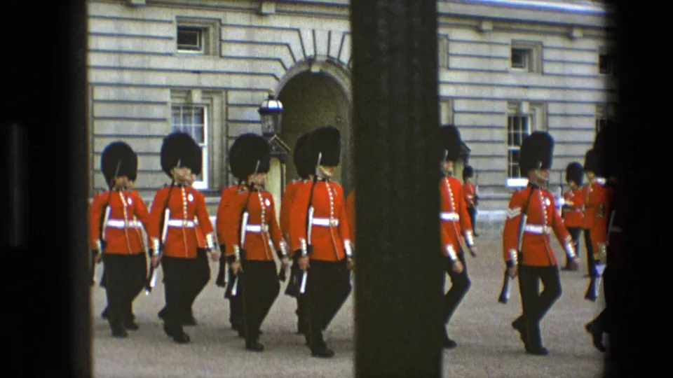 1969 England home-movie still showing several Buckingham Palace guards in red tunics walking in formation behind a dark foreground bar.