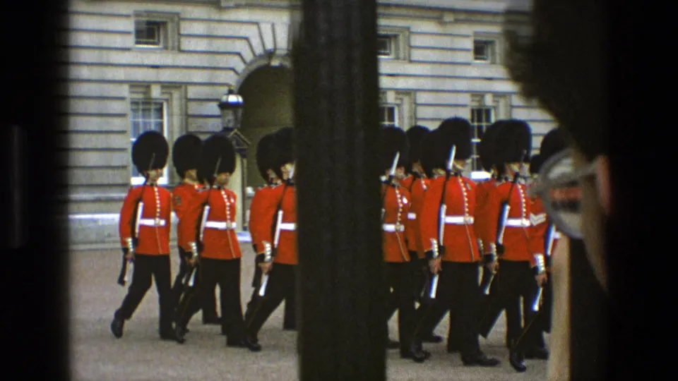 1969 England frame with Buckingham Palace guards in ceremonial red uniforms marching past stone architecture, partly blocked by railings.