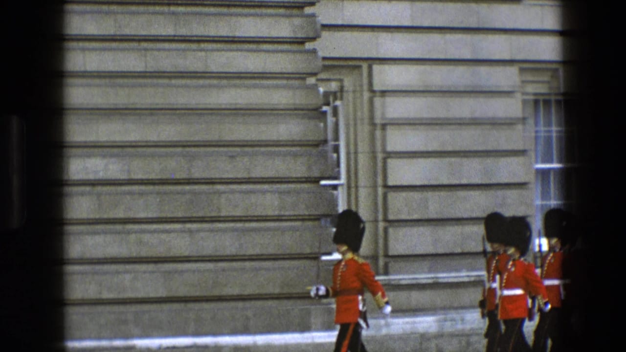 British guards in red tunics march past a grey stone palace wall, partly blocked by dark foreground bars.