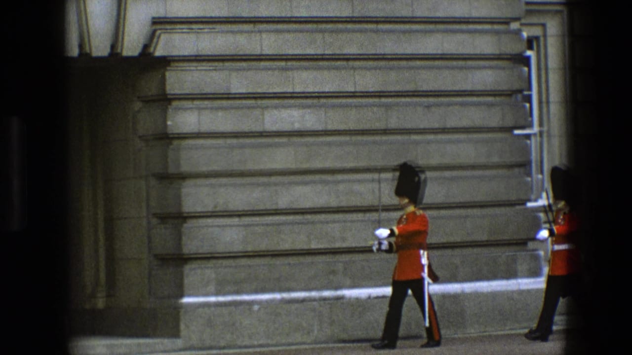 A guard in a red tunic and bearskin hat marches along a stone building, with another guard partly visible.