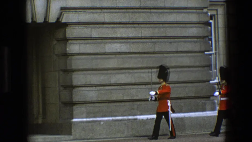1969 England frame of a Royal Guard marching in profile beside palace stonework, with a second uniformed guard at the edge.