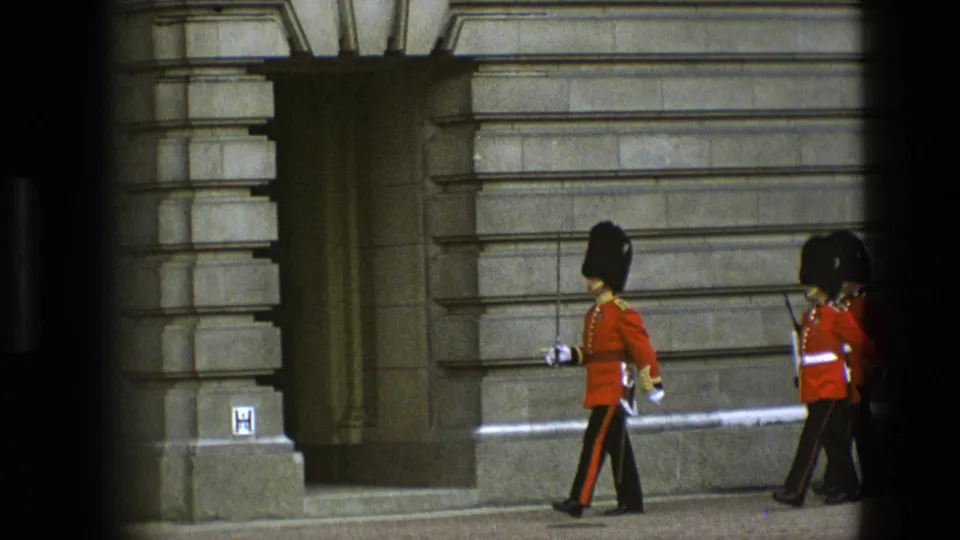 1969 England home-movie view of two Royal Guards marching past grey stone palace architecture, partly obscured by foreground bars.