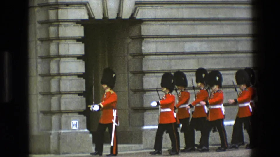 1969 England frame showing a small formation of Royal Guards in red tunics and bearskin hats marching beside palace stonework.