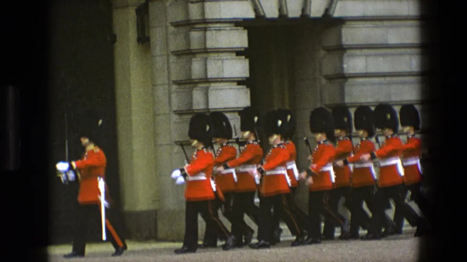 Ceremonial guards in red tunics and black hats move in synchronized formation outside a palace-like stone exterior.