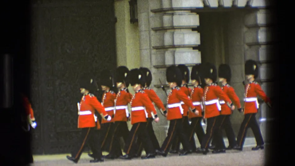 A longer line of British ceremonial guards marches left to right in front of a grand stone palace wall.