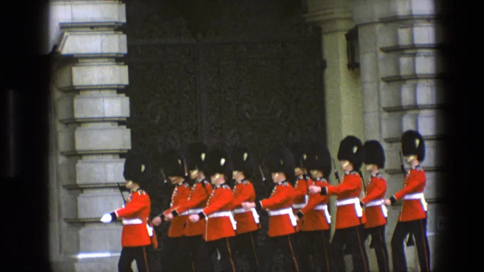 British ceremonial guards in red dress uniforms continue their march beside palace architecture in 1969 England footage.