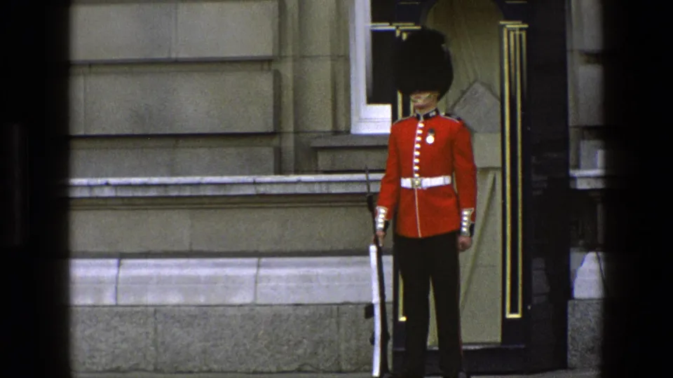 A Queen's Guard stands still at a Buckingham Palace doorway in 1969 England footage, wearing red tunic, bearskin hat, and white belt.