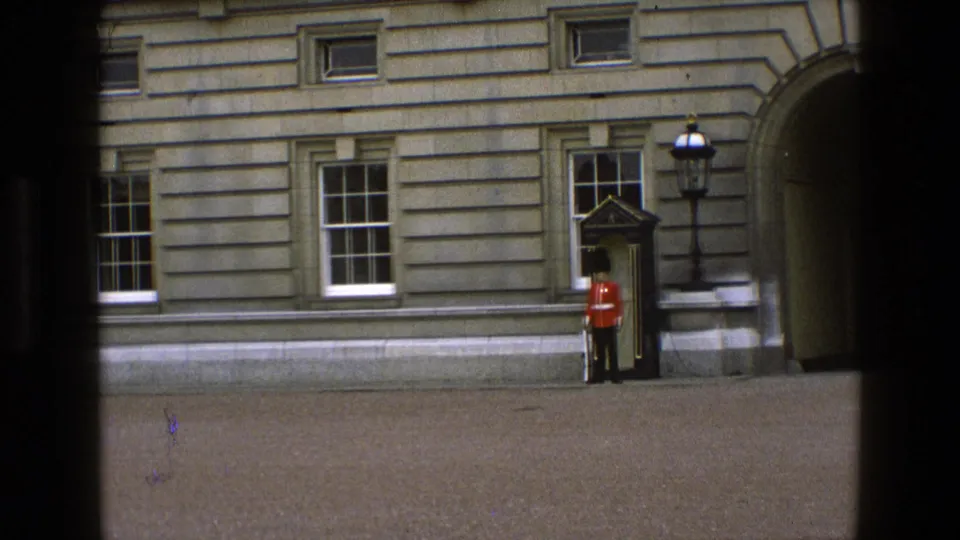 1969 England 8mm frame with a Royal Guard in red uniform posted near a sentry box, windows, lamp, and arched doorway.