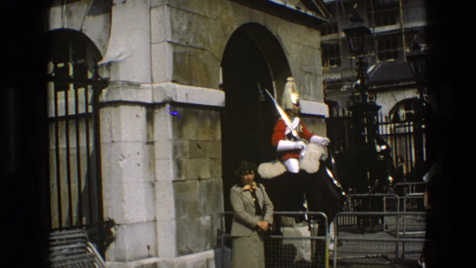 1969 England home-movie frame showing a mounted ceremonial guard at a stone gate while a woman stands nearby.