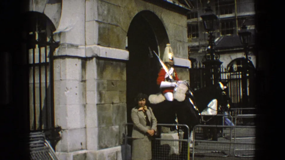 1969 England frame of a ceremonial rider on a black horse near iron gates, with a civilian woman in view.