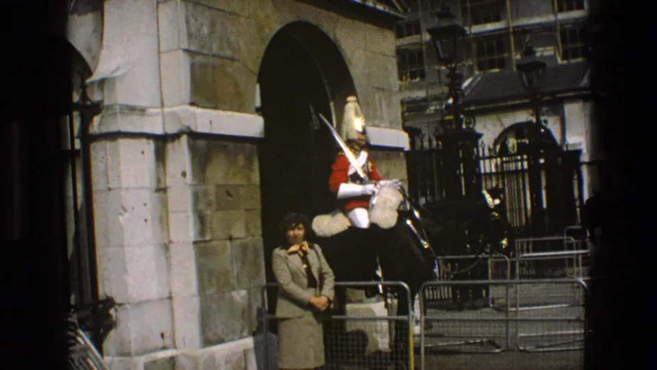 1969 England home-movie view of a mounted guard under a stone archway, framed by barriers and a civilian observer.