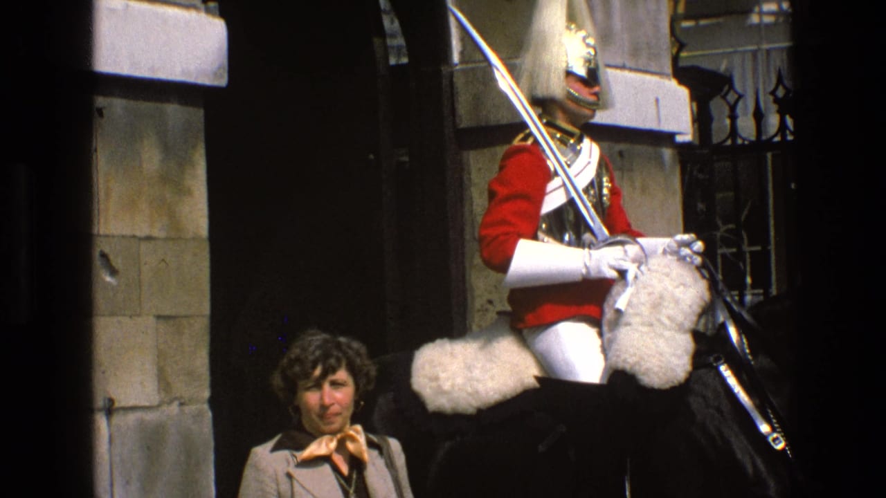 A woman faces the camera while a mounted ceremonial guard in red uniform sits on a black horse beside stonework in England, 1969.