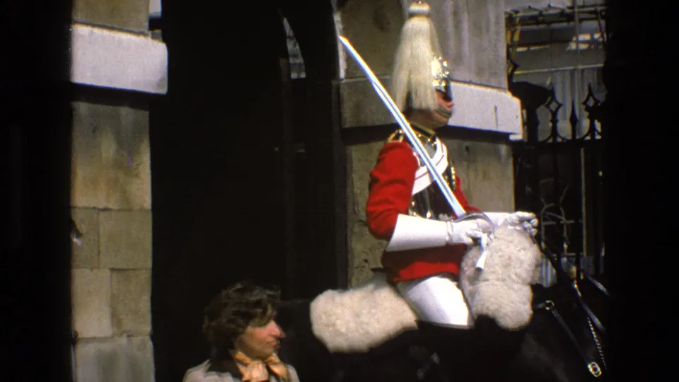 At the end of the clip, the woman turns sideways while the mounted guard remains seated with sword visible near stonework and fencing.