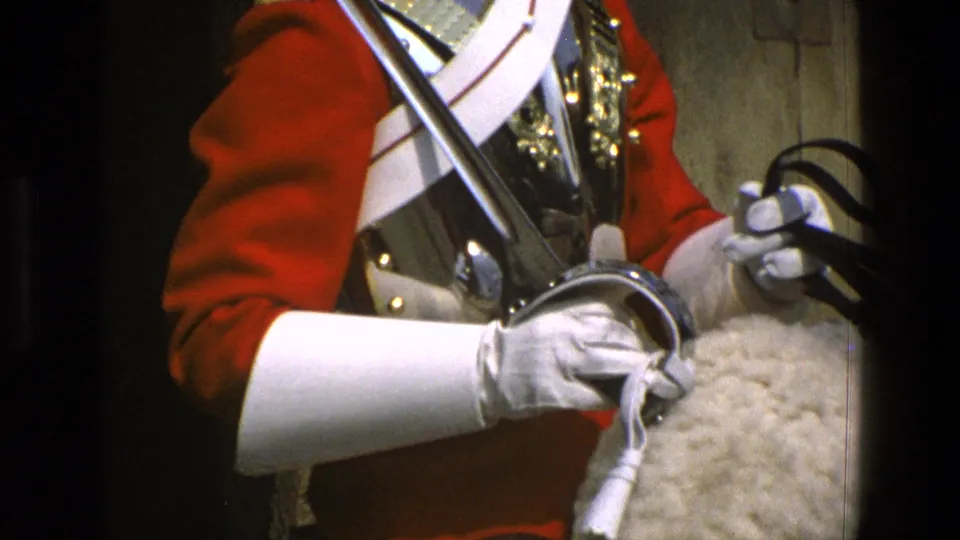 Tight 8mm frame of a mounted ceremonial guard in red and white uniform holding reins over a pale saddle cover in England.