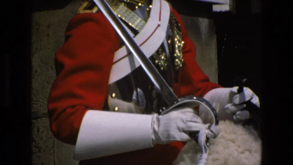 Close 1969 England frame showing a mounted ceremonial guard's red tunic, metal cuirass, sword, white gloves and reins on horseback.