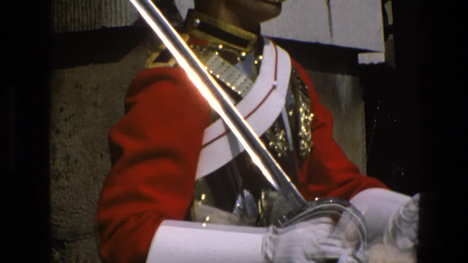 1969 England home-movie frame showing a ceremonial guard's red tunic, white gloves, armor, cross belt, and angled saber.