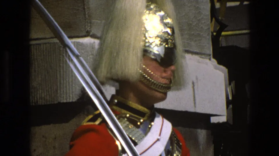1969 England home-movie close-up of the ceremonial guard's plumed helmet, angled saber, red uniform, and white cross belt.