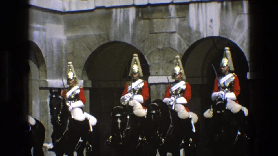 1969 England 8mm frame showing mounted ceremonial riders in red coats and plumed helmets lined up before arched stone doorways.