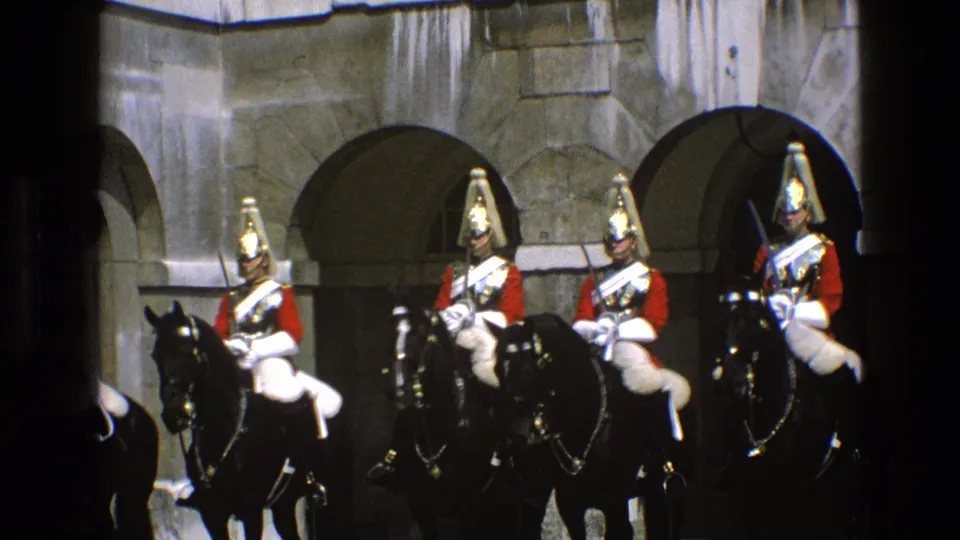 England 1969 home-movie frame with mounted ceremonial riders in red and white uniforms seated on black horses near stone archways.