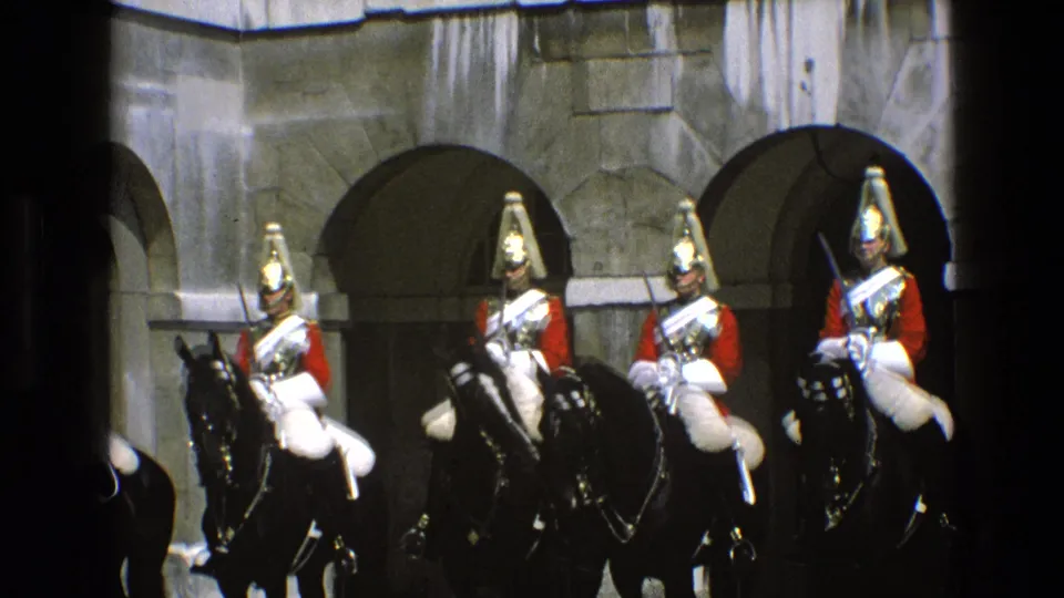 Four mounted guards in ceremonial dress appear on black horses in 1969 England footage, with arched stone architecture behind them.