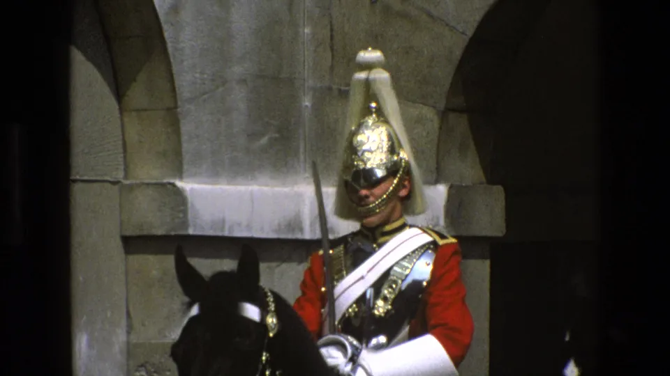Mounted guard in ceremonial uniform faces forward on a black horse, with a sword held upright against stone architecture.