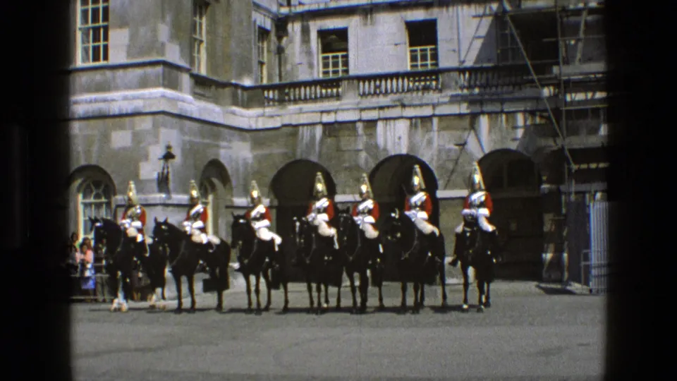 1969 England home-movie frame showing mounted ceremonial guards in red tunics before a grand stone building with arched openings.