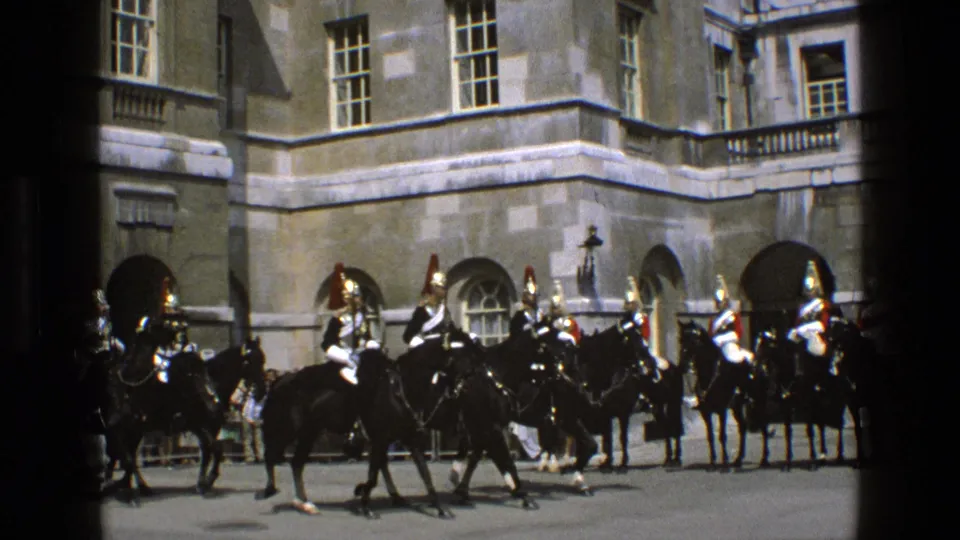 1969 England home-movie still showing a mounted guard crossing before a line of ceremonial riders outside a stone palace-like building.