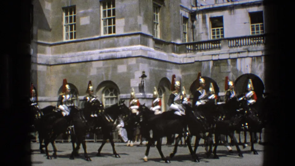 1969 England 8mm frame of mounted guards in plumed helmets riding black horses before arched palace windows.