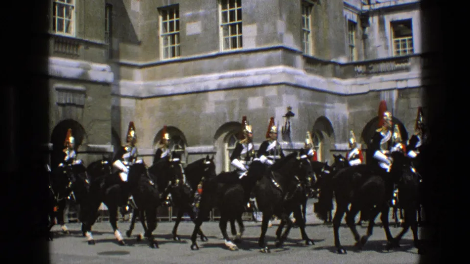 Home-movie frame of mounted guards in ornate helmets riding black horses before a sunlit palace facade in England.