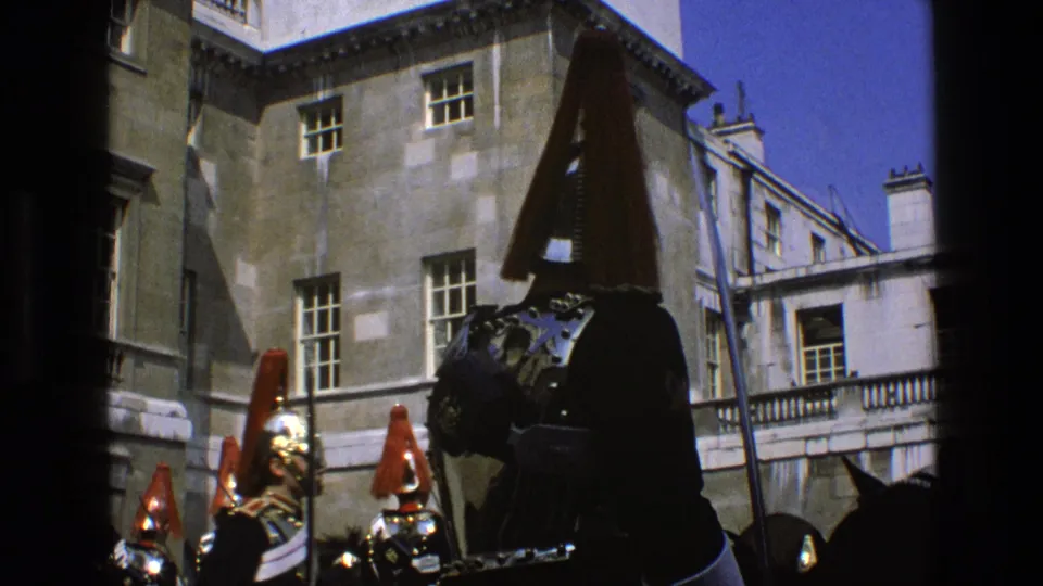 1969 England 8mm frame showing ceremonial mounted guards with red plumes and reflective armor near a formal stone courtyard.