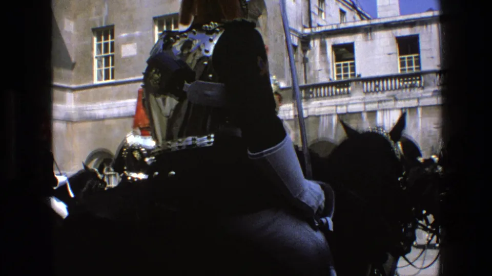 Rear view of a ceremonial mounted guard in England, with red-plumed helmets and stone architecture visible on 8mm film.