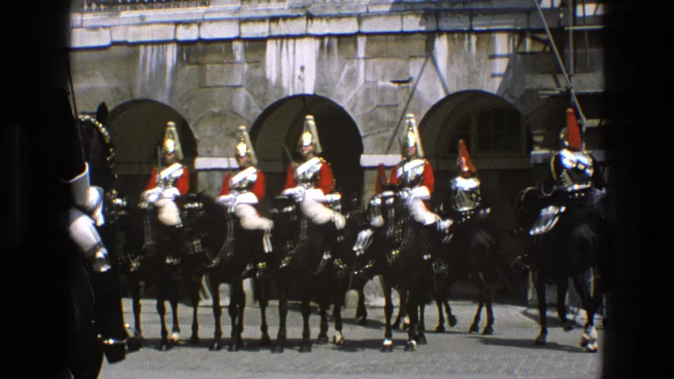1969 England home-movie frame with mounted ceremonial soldiers and horses filling the courtyard view.