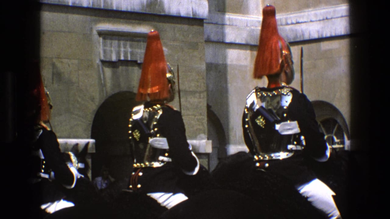 Mounted guards in red plumes and dark uniforms seen from behind near a stone building.