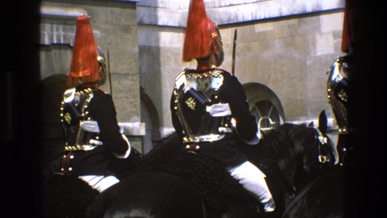 1969 England home-movie frame of mounted guards in ornate ceremonial uniforms, viewed from behind against a stone facade.