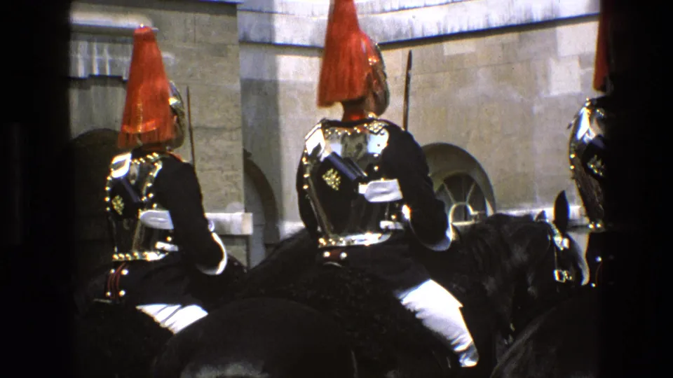 1969 England home-movie frame of mounted guards in ornate ceremonial uniforms, viewed from behind against a stone facade.